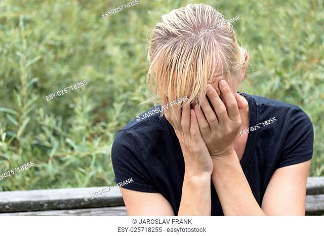 Desperate blonde middle aged woman in black shirt is sitting on the bench outside. Hands are covering her face