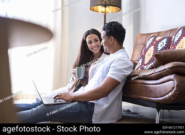 Interracial couple boy and girl use laptop computer at home sitting on the floor against sofa and looking each other with a smile - happy young people in home...