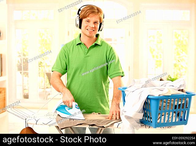 Portrait of young goodlooking man with headphones ironing clothes in living room, looking at camera, smiling