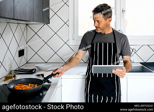 Man cooking stir-fried vegetables in pan holding tablet PC at home