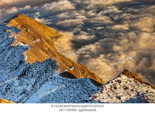 Tussock covered ridge below Mt Roy partly covered by winter snow, above sea of cloud over Lake Wanaka, Central Otago, Southern Alps, New Zealand