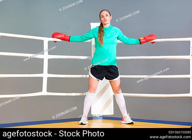 Confident beautiful young athlete woman with collected hair standing in the corner of boxing ring in boxing red gloves and looking at camera