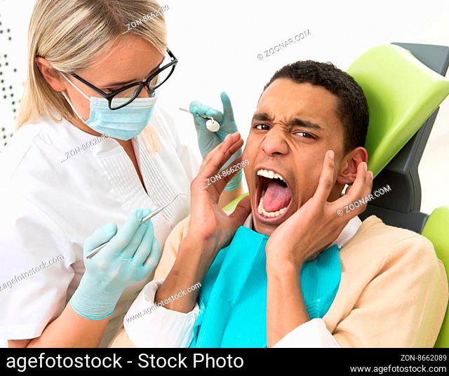 Young man screaming and shouting at dentist#39;s office while young dentist lady having his teeth examination