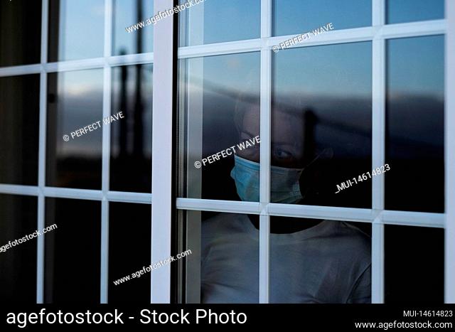close up of infected woman in quarentine at home or hospital looking out of the window - sad and bored woman looking away - wearing medical mask to prevent any...