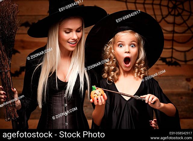 Halloween Concept: beautiful caucasian mother and her daughter in witch costumes enjoy using magic with magic wand to halloween pumpkin jar over bats and spider...