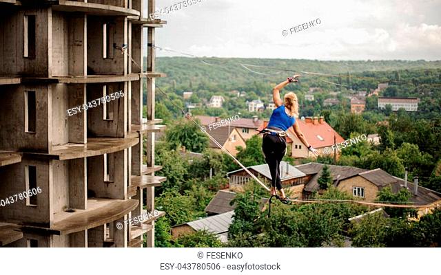 Rear view blondie girl walking on the slackline rope on the background of houses among trees and clear sky