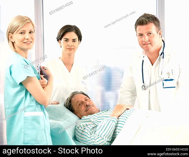 Portrait of senior patient lying in bed, surrounded with hospital crew, smiling at camera