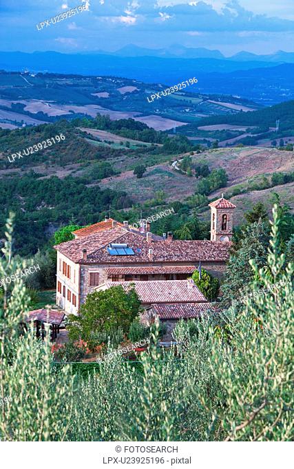 Aerial view of borgo farmhouse with tiled rooftops and church bell tower, fields, hills and distant mountains beyond, sunny late afternoon, Umbria, Italy