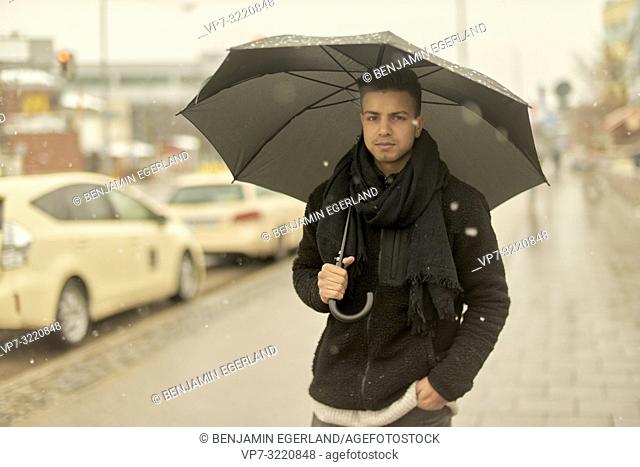 young man, Afghan ethnicity, with umbrella walking outdoors in city during winter, snowing, in Munich, Germany