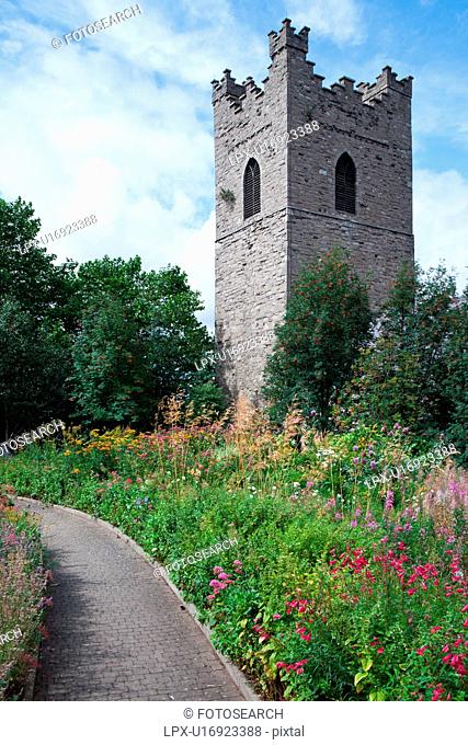 Romanesque square tower of St. Auden's Church on sunny summer afternoon, path through colourful flowerbeds, blue sky, Dublin, Southern Ireland