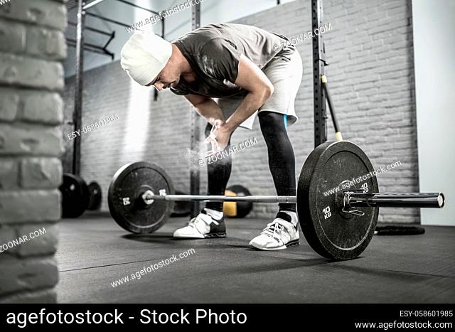 Healthy guy with a beard tilts to a barbell in the gym on the gray brick wall background. He wears sportswear, white sneakers and a white cap