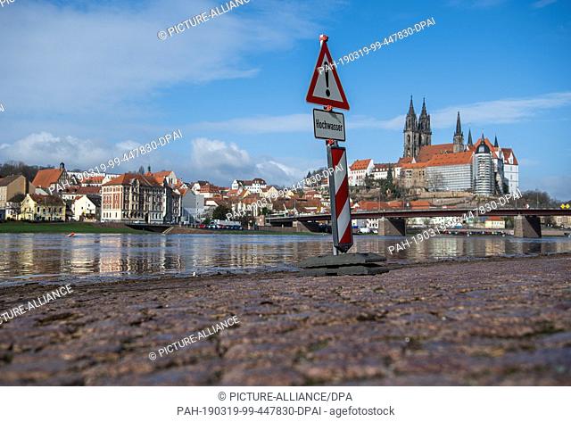 19 March 2019, Saxony, Meißen: A sign on the Elbe parking lot warns of flooding, in the background you can see Albrechtsburg Castle with the cathedral