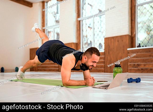 Handsome young sport man doing one leg up push ups exercise in empty gym or home watching online sports videos all in black clothes