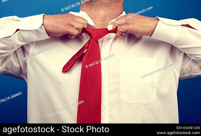 adult man in a white office shirt tears off a red satin tie from his neck on a blue background, concept of depression, unemployment and dismissal