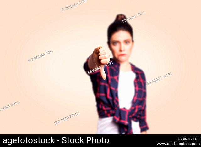 Dislike. Young unhappy upset girl with casual style and bun hair thumbs down her finger, on beige blank wall with copy space looking at camera with toothy smile