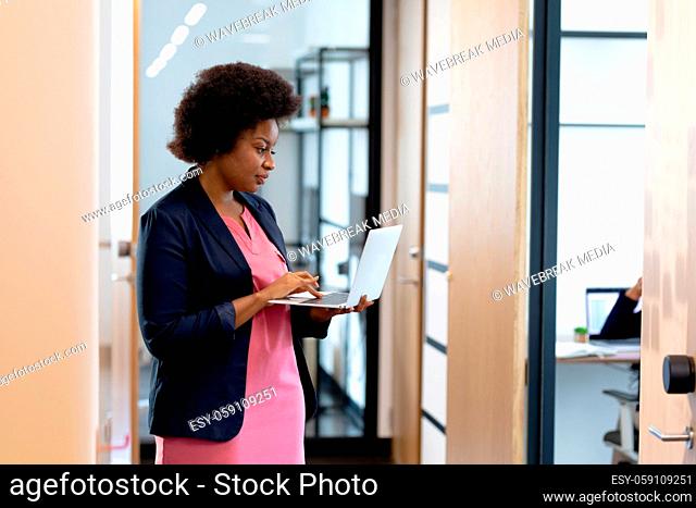 Smartly dressed african american businesswoman standing in corridor using laptop