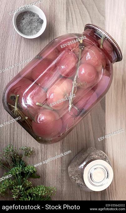 Home preservation: a large glass jar with red ripe pickled tomatoes, closed with a metal lid, next to spices and herbs. Top view, close-up