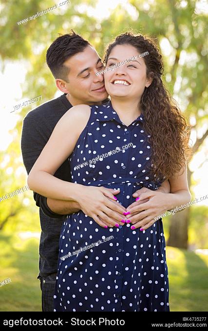 Happy hispanic man hugs his pregnant wife outdoors at the park