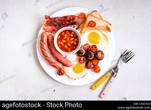 Full english breakfast with fried eggs, tomatoes, sausages, bacon, mushrooms, toasts and beans. Breakfast on a white plate with forks on the white wooden table