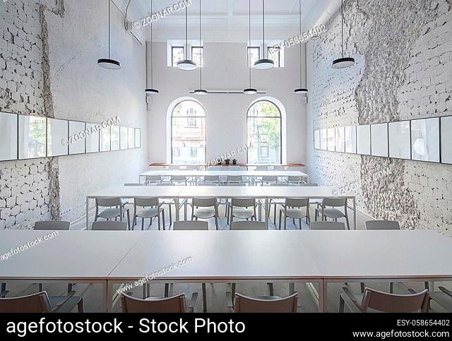 Nice hall in a cafe with shabby light walls and gray floor. There are white tables with chairs, wooden rack with plants in the pots, arch windows