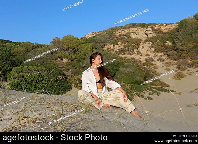 Thoughtful young woman sitting on sand at beach in front of blue sky, Patara, Turkiye