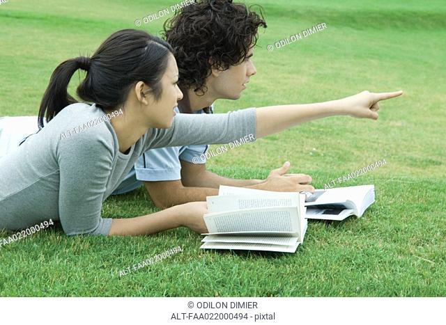 Young couple lying on grass with books, woman pointing, both looking out of frame