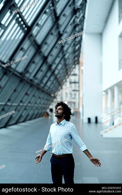 Male entrepreneur standing with arms outstretched looking up in corridor at office