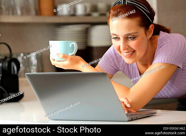 Happy woman using laptop and holding coffee cup in the kitchen at home