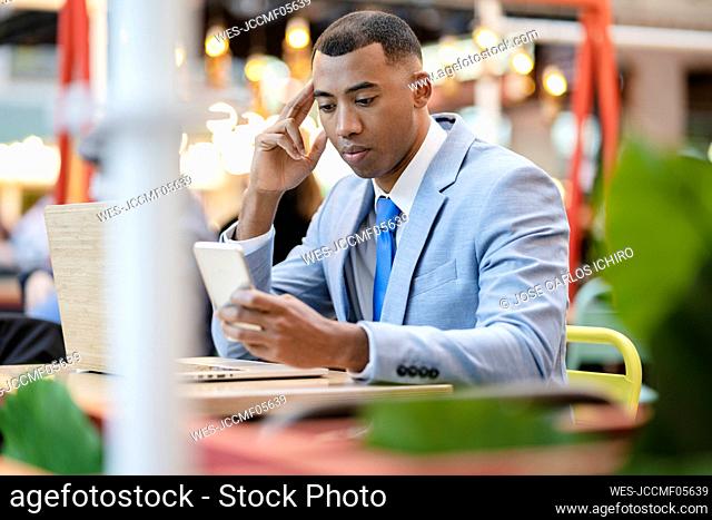 Businessman using smart phone sitting with laptop at sidewalk cafe