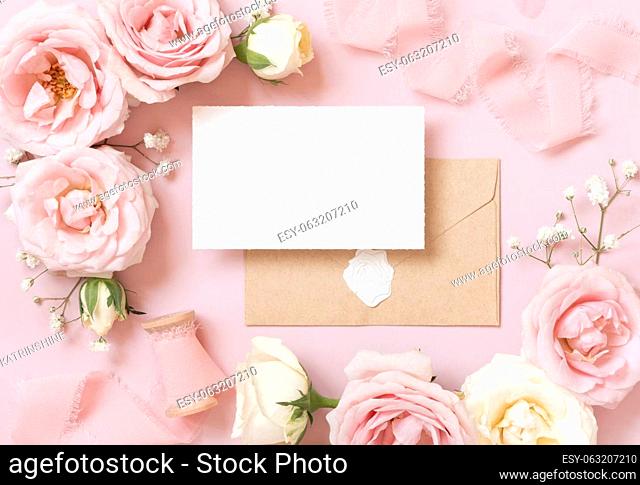 Card with envelope between light pink roses and silk ribbons on pink top view, wedding mockup. Romantic scene with vertical blank card and pastel flowers flat...