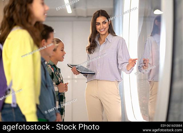 Information. Smiling woman holding clipboard pointing hand to side and students standing interested looking in hall indoors