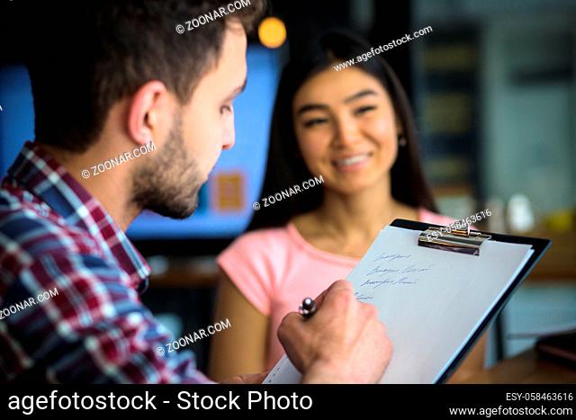 Picture of beautiful lady having interview in restaurant or cafe. Cute man writing answers to questions on clipboard