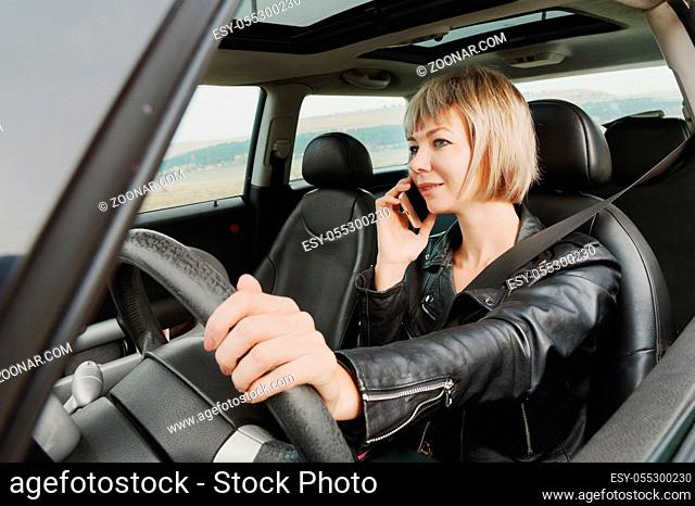 Business girl in leather jacket at the wheel of a car is wearing a seat belt talking on the phone