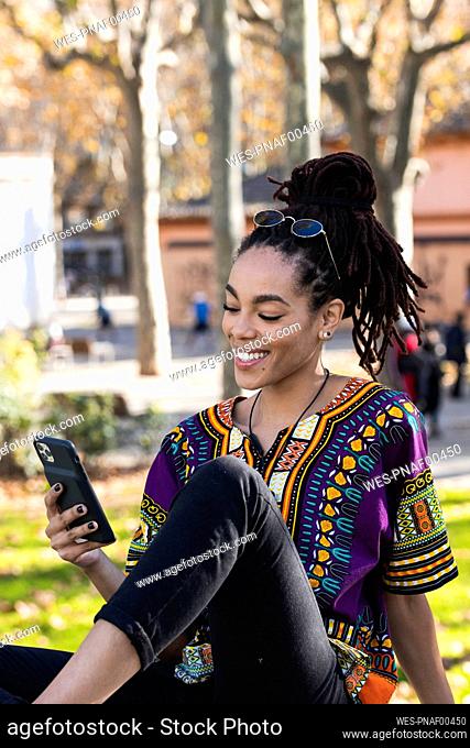 Smiling fashionable young woman using smart phone while sitting at park