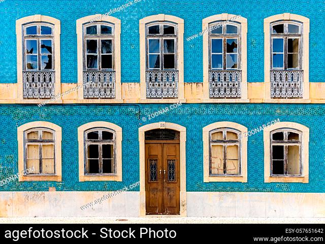 A frontal view of a house facade covered in azulejo tiles with broken windows and wooden door