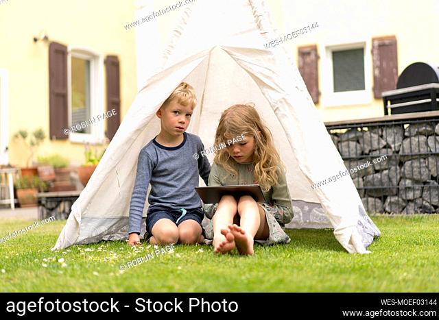 Cute siblings with digital tablet sitting in tent on grass at back yard against house