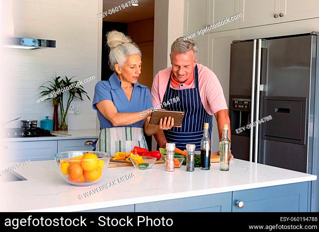 Happy caucasian senior couple standing in kitchen, using tablet and preparing meal together