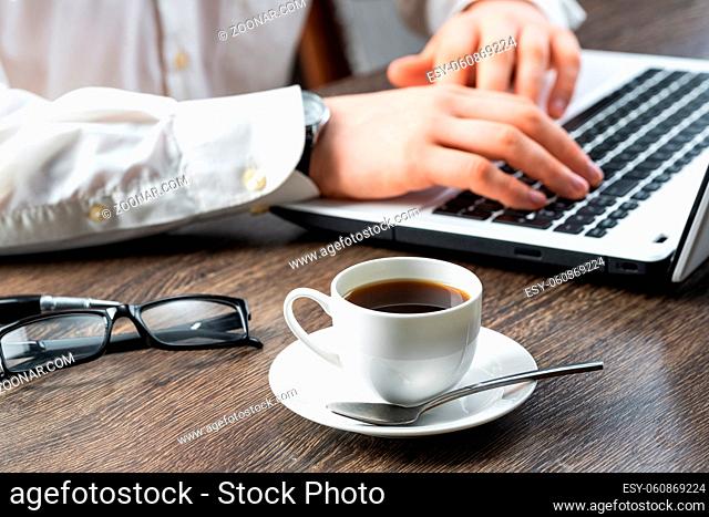 Businessman sitting at desk and using laptop. Close-up of man hands typing on keyboard in office. Side view consultation workplace with glasses and coffee cup