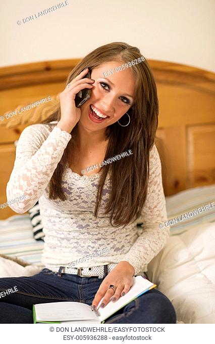 Teenage girl lying on a bed talking on a cell phone while doing homework