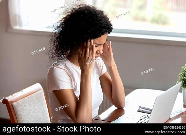 Tired stressed mixed race woman sitting at table near laptop touch massaging temples suffers from headache overuse of computer african 30s female feeling pain...