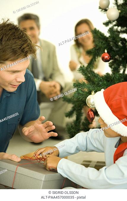 Father and son opening Christmas present in front of Christmas tree, boy wearing Santa hat
