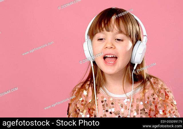 Pretty 6 or 7 years old little girl singing and dancing with headphones. While posing on pink background with copy space in studio