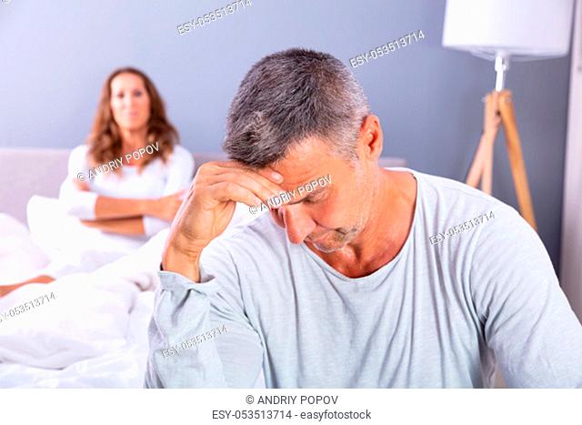Close-up Of Depressed Man Sitting On Bed In Front Of Her Wife At Home