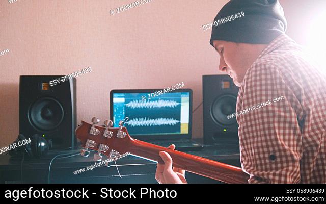 Young man in hat musician composes and records soundtrack playing the guitar using computer, headphones and keyboard, focus on equipment
