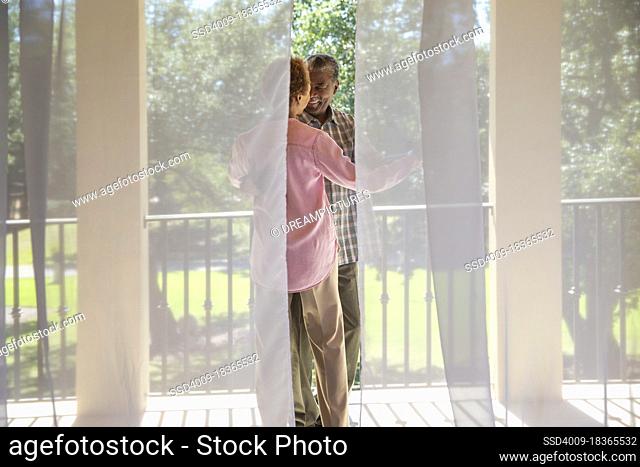 Older couple dancing on porch overlooking green lawn , seen through sheer curtains