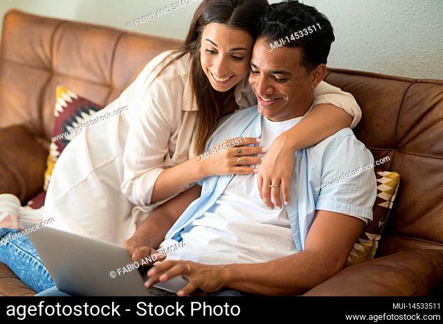 Happy interracial caucasian and black couple with young woman hugging her boyfriend while he works on laptop. Relationship and friendship in smart working job...