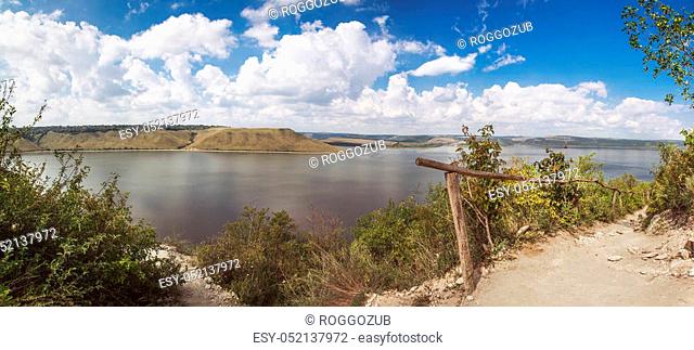 lake shore with forest under sky. Trees on rock coast of rippling lake in sunny day. Beautiful panoramic landscape with trees on big stones and lake