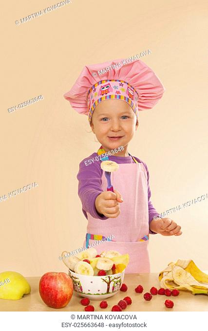 Little girl in purple cook hat and tablier making fruit salad. Healthy eating habits
