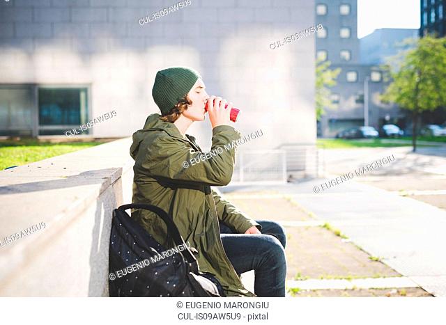 Young man sitting on urban wall drinking from can
