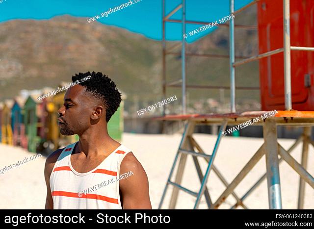 African american young man looking away while standing at beach against mountain on sunny day
